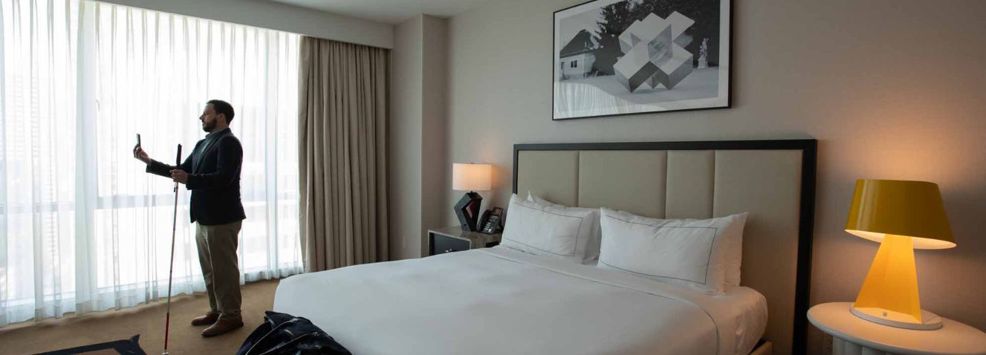 Description provided by Be My AI: Hotel room interior with a large, neatly made bed in the center, featuring white linens and multiple pillows. To the right, on a round bedside table, sits a modern yellow lamp that is turned on. Above the bed, there is a framed black-and-white artwork or photograph on the wall. In the left section of the image, a man with a beard stands near floor-to-ceiling curtains, holding a white cane and using a smartphone, facing the light streaming through the window. The room has a clean, modern feel with neutral-colored walls, carpeting, and a nightstand with another lamp and a telephone. A piece of dark clothing appears draped at the foot of the bed.