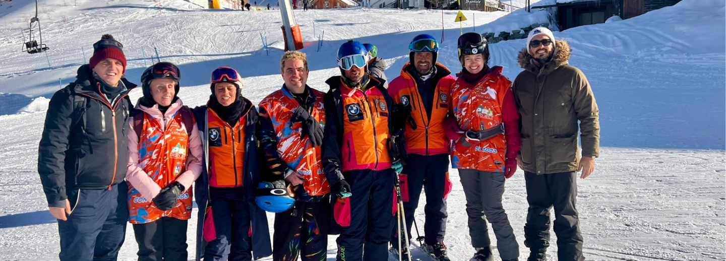 A group of eight people standing together on a snowy ski slope. Most of them are wearing colorful orange and red jackets, some with helmets and ski goggles, suggesting they are part of a ski or winter sports team. A few people are dressed in darker winter coats and hats. Some are holding ski poles, and there is snow all around with ski lifts and equipment visible in the background. The group looks happy and is posing for the photo in bright daylight.